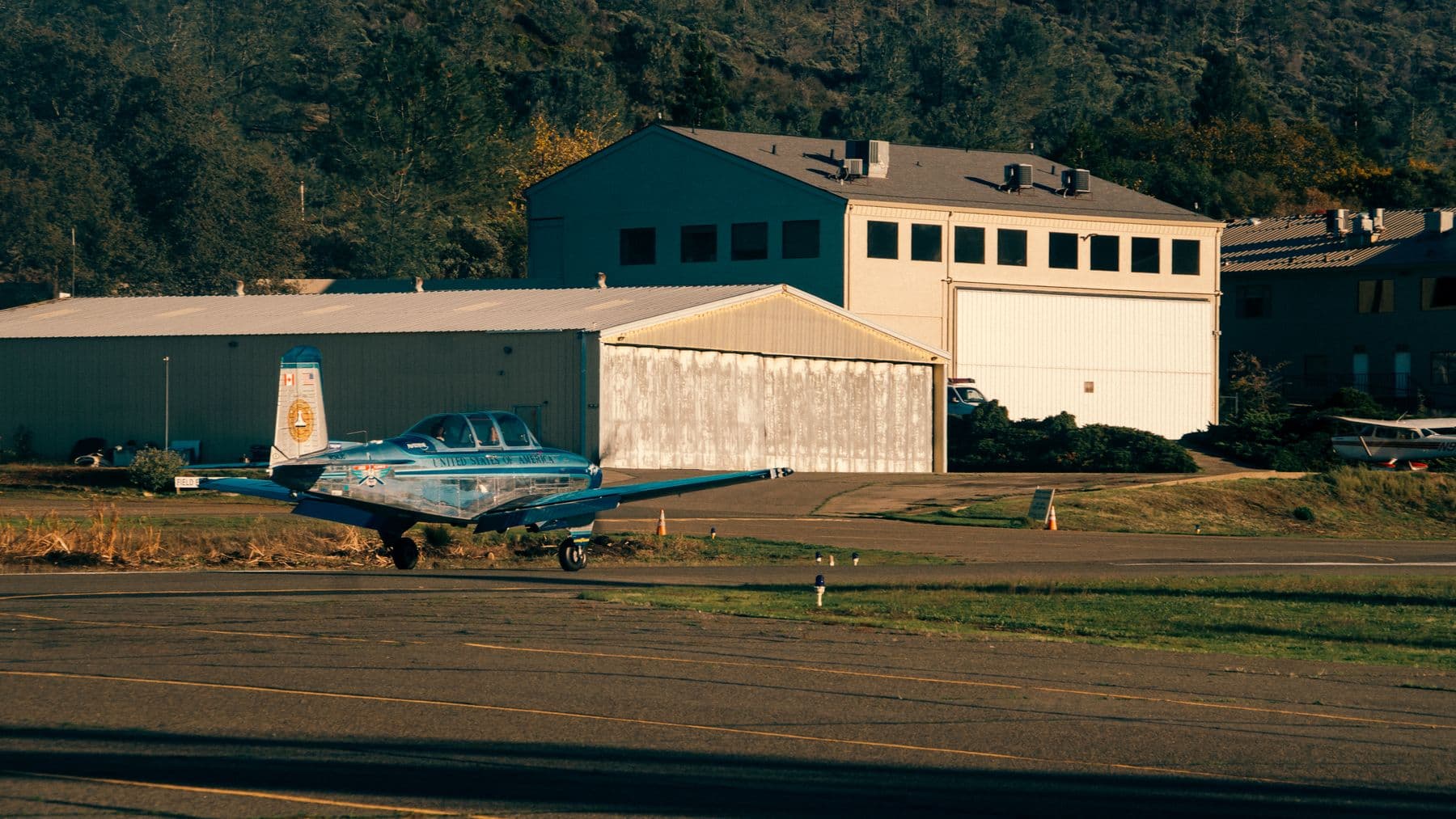 A small blue propeller plane parked on a grass-edged airfield in front of old hangars.