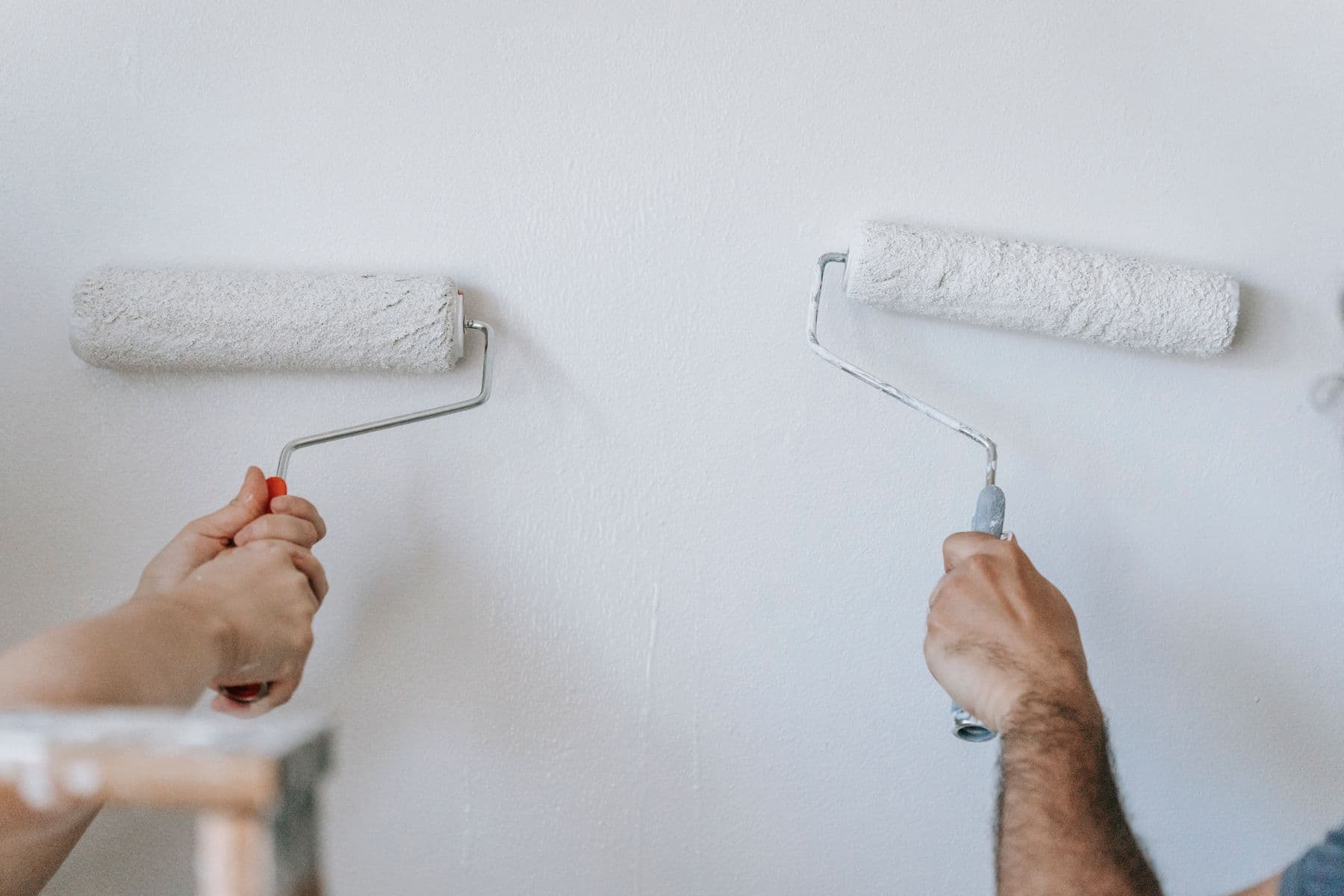 Two hands rolling white paint onto a white wall with paint rollers.