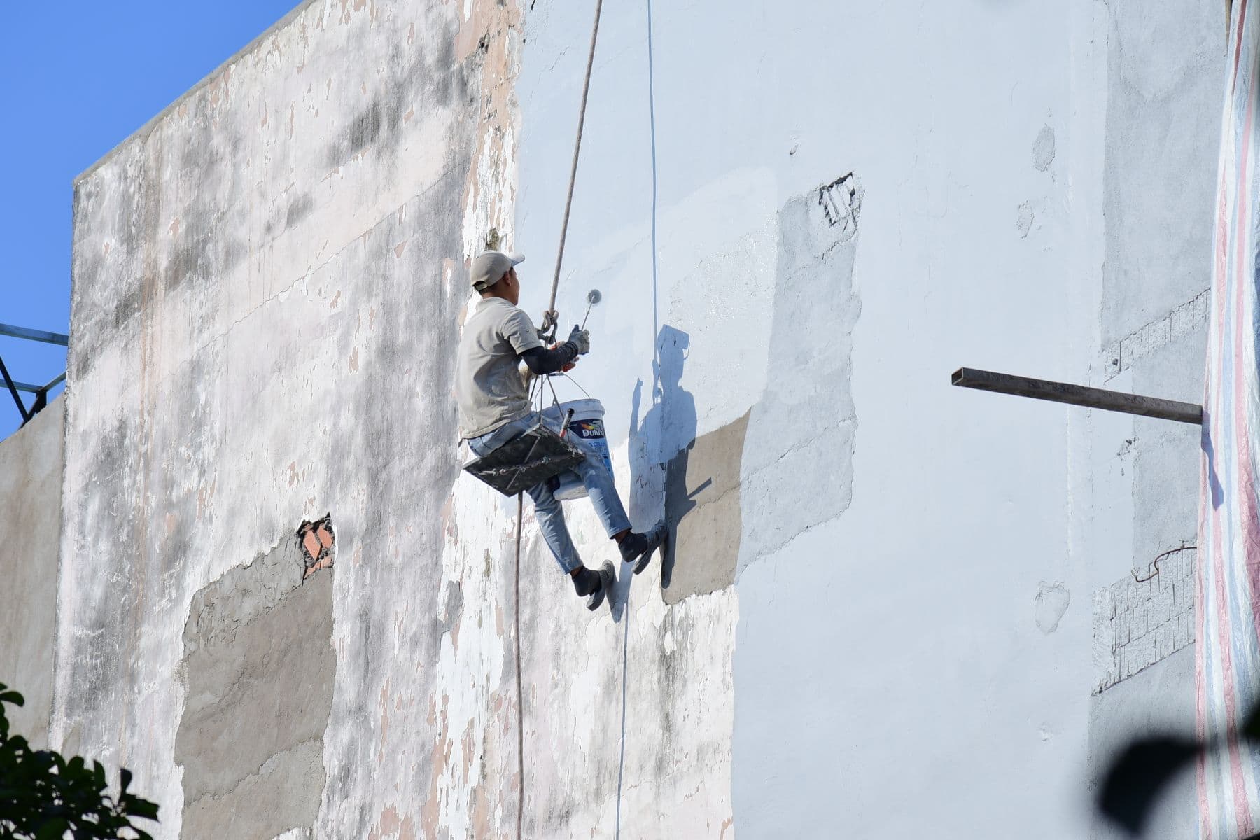 A rope-access painter rolling white paint onto a stained concrete wall.