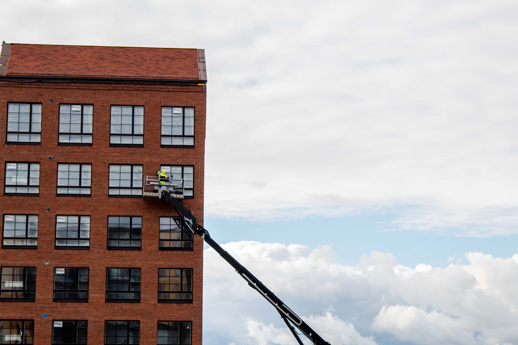A worker on a telescopic lift at an upper window of a red brick building, with clouds behind.