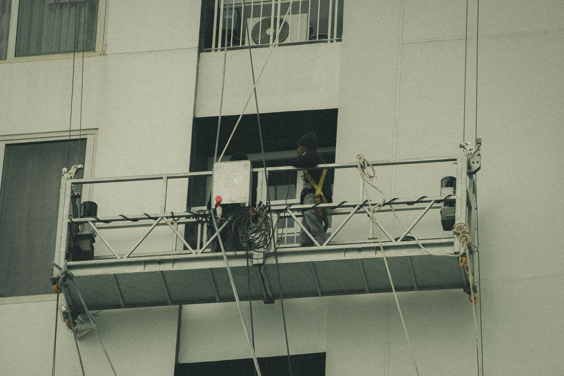 A worker on a suspended platform hanging against the pale wall of a building.