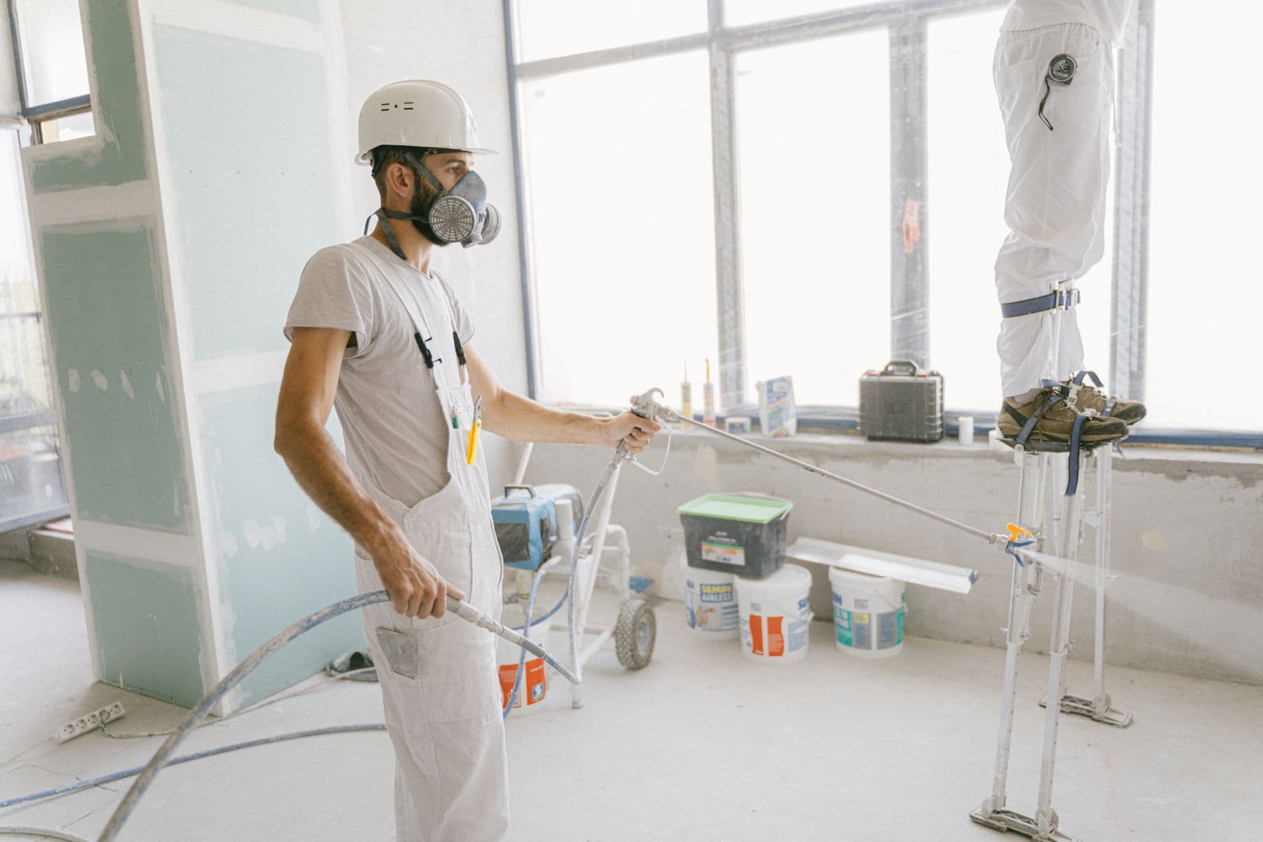 A worker in a hard hat and respirator spraying paint on an interior wall with an airless sprayer.